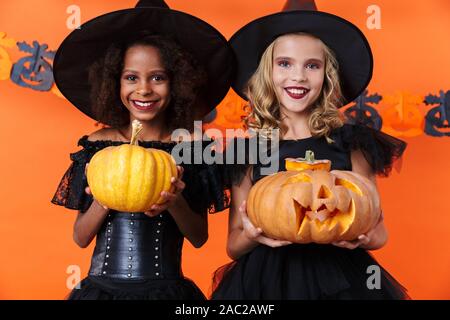 Bild des freudigen multinationale Mädchen in Schwarz halloween Kostüme Kürbisse Holding und lächelnd über orange Wand isoliert Stockfoto