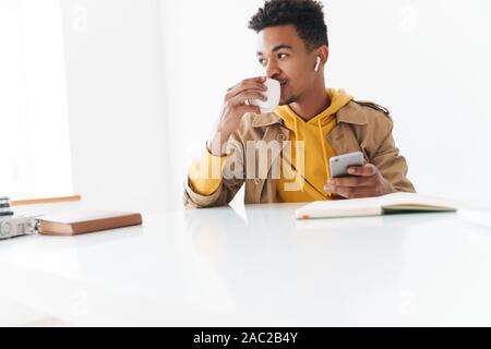 Bild von optimistisch african american Guy holding Smartphone und Kaffee Tasse beim Sitzen am Tisch in Weiß Büro Stockfoto
