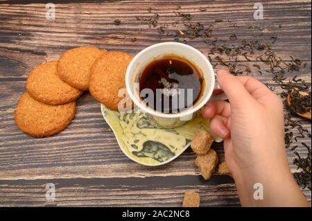 Die Hand des Mädchens hält eine Tasse schwarzen Tee, oatmeal Cookies, Tee Blätter, brauner Zucker auf einer hölzernen Hintergrund. Nahaufnahme Stockfoto