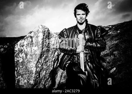 Portrait von stattlichen Ritter in Kostüm neben Rock holding Schwert und Kamera Stockfoto