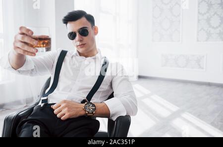 Lassen Sie uns einen Schuß. Junge stilvolle Geschäftsmann in Sonnenbrille sitzt auf einem schwarzen Stuhl mit Glas Whiskey Stockfoto