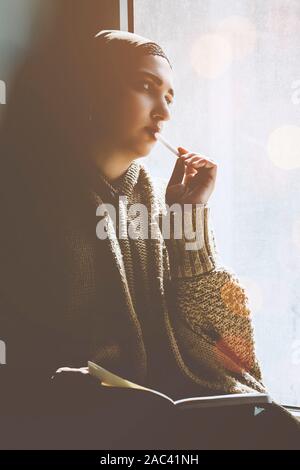 Junge muslimische Frau sitzt auf der Fensterbank. Mitte - östliche Verfasser Frau nachdenklich sitzen in der Nähe der Fenster und ihre Geschichte schreiben auf dem Merkzettel. Ira Stockfoto