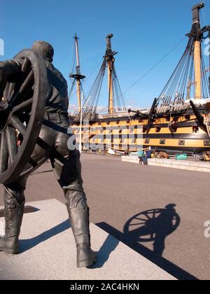 AJAXNETPHOTO. PORTSMOUTH, England. - HISTORIC DOCKYARD BRONZESTATUE - EIN FELD GUNNER dargestellt anheben EIN FELD GUN RAD von britischen Bildhauers Christopher Kelly. Die STATUE AUF EINEM SOCKEL ERINNERT AN MÄNNER VON PORTSMOUTH BEFEHL IN DER JÄHRLICHEN FIELD GUN WETTBEWERB AN DER ROYAL TURNIER UND AN DIE ROYAL NAVAL KASERNE VON 1947-1999 geschult, wenn das Turnier beendet. Bis AN DIE GRENZE UND DARÜBER HINAUS. NELSON'S 1805 Schlacht von Trafalgar FLAGGSCHIFF HMS VICTORY wird gesehen, Hintergrund. Foto: Jonathan Eastland/AJAX REF: GR 110111 3170 Stockfoto