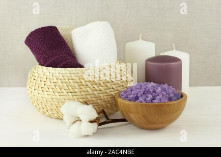Lavender Meersalz für Badewanne in einem Bambus Schale, Handtücher aus Baumwolle in einer korbwaren Vase und Kerzen auf einem weißen Tisch gegen eine graue Wand stand Stockfoto