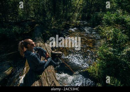 Schöne Mädchen sitzt auf Anmelden und Chillen nach Reisen. Frau Reisende im Sommer geht auf hölzernen Brücke im Hintergrund von Wald. Bridged liegt auf der Ri Stockfoto