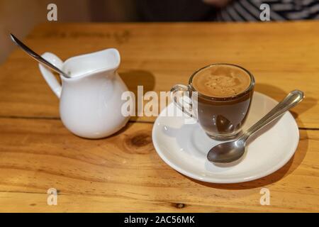 Glas Becher Kaffee mit Löffel und Milch auf weißem Teller Stockfoto