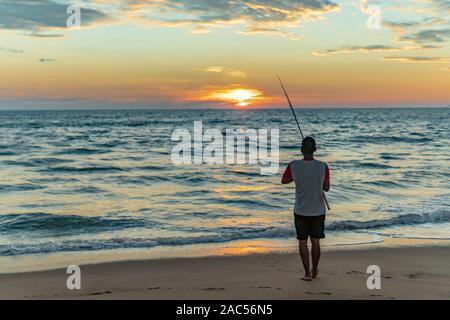 Mann mit Angelrute steht auf sandigen Strand bei Sonnenuntergang in Goa, Indien Stockfoto