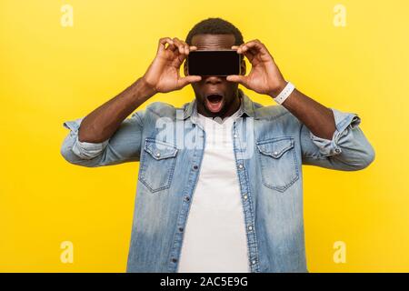 Portrait von fassungslos erstaunt Mann in Jeans Hemd, die Augen mit Handy, ausblenden Gesicht anonym zu sein, mit Gadget mit Wow schockiert Ausdruck. ind Stockfoto