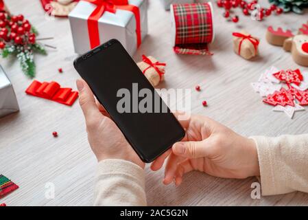 Mobiltelefon Mockup in der Frau die Hände. Weihnachten und neues Jahr Dekorationen und Geschenke im Hintergrund. Close-up. Stockfoto