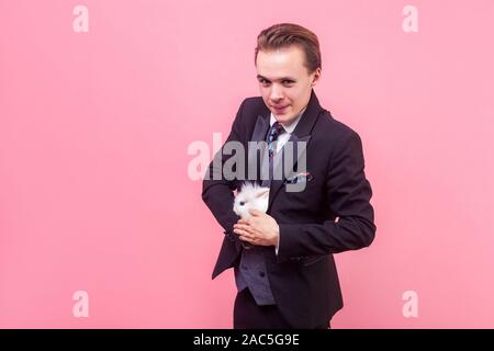 Portrait von lustigen positiven Mann im eleganten Anzug und mit stilvollen Frisur verstecken weißen Kaninchen in seinem Smoking, als ob Zauberer, Trick, grinsend an Kameras Stockfoto