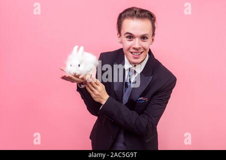 Pet care. Portrait von fröhlichen gutaussehenden Mann im eleganten Anzug und mit stilvollen Frisur ständigen Holding cute bunny Hase auf seine Handfläche, in die Kamera lächeln. Stockfoto