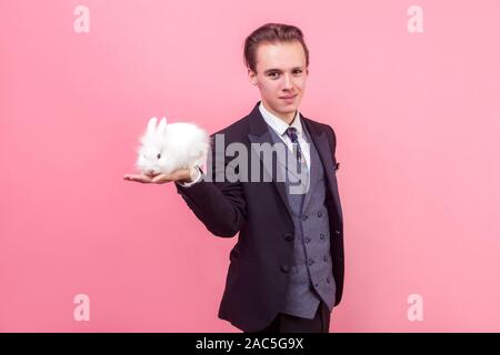 Pet care. Porträt der jungen Hübschen positiver Mann im eleganten Anzug mit modischen Frisur ständigen Holding cute bunny Hase auf seinem Palm, lächelnd an Kameras Stockfoto