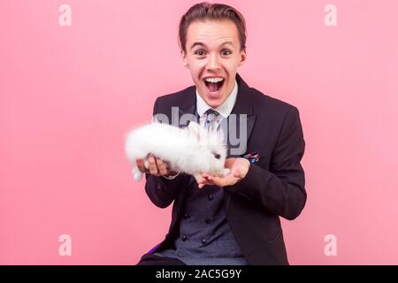 Pet Adoption. Portrait von extrem aufgeregt, Mann im eleganten Anzug mit modischen Frisur Holding niedliche Kaninchen auf seinem Palm, an Kamera suchen mit Erstaunen Stockfoto