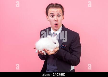 Pet Adoption. Portrait von extrem aufgeregt, Mann im eleganten Anzug mit modischen Frisur Holding niedliche Kaninchen auf seinem Palm, an Kamera suchen mit Erstaunen Stockfoto