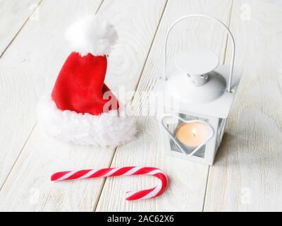 Santa claus red hat, weiß Laterne mit einer brennenden Kerze und süße Candy Cane auf weißem Holz- Oberfläche. Weihnachten und Neujahr Grußkarte. Stockfoto