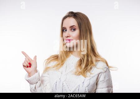 Attraktive junge blonde Frau in Beige gestrickter Pullover und Finger auf Kopie Raum zeigt auf weißem Hintergrund. Produkt Präsentation, Werbung, Stockfoto