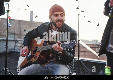 Junger bärtiger Mann. Kerl mit akustischer Gitarre singt. Freunde haben Spaß an der Party auf dem Dach mit dekorativen farbigen Glühlampen Stockfoto