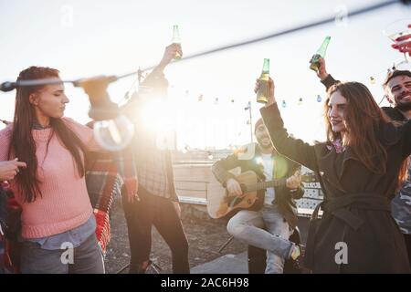 Tanzen in der Sonne. Gruppe von jungen Menschen, die fest an eine Dachterrasse mit etwas Alkohol und Gitarre spielen Stockfoto