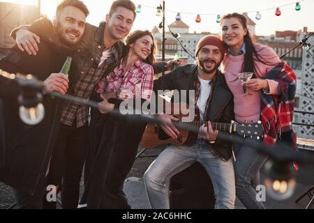 Schöne junge Menschen. Party auf dem Dach. Fünf gute Freunde suchen, die für das Bild mit Alkohol und Gitarre posieren Stockfoto