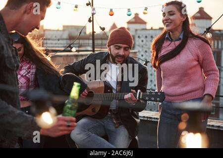 Spielen auf dem Instrument ist immer gut zu haben. Gitarrist sitzt und singt für seine Freunde auf der Dachterrasse mit dekorativen farbiges Licht Stockfoto