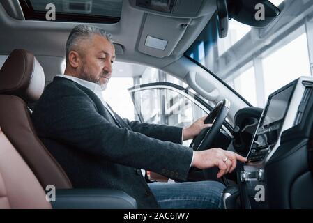 Senior Geschäftsmann in der offiziellen Kleidung sitzt in einem Luxus Auto und Drücken der Tasten auf der Music Player Stockfoto