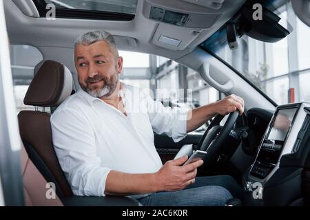 Lächelnd, während in einem ganz neuen Auto sitzen. Bärtige älterer Mann im weißen Hemd hält Mobiltelefon Stockfoto