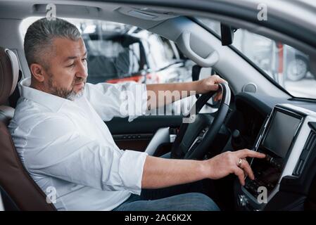 Alles perfekt funktioniert. Senior Geschäftsmann in der offiziellen Kleidung sitzt in einem Luxus Auto und Drücken der Tasten auf der Music Player Stockfoto