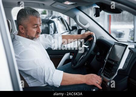 Kaufen und Testen neuer Automobile. Senior Geschäftsmann in der offiziellen Kleidung sitzt in einem Luxus Auto und Drücken der Tasten auf der Music Player Stockfoto