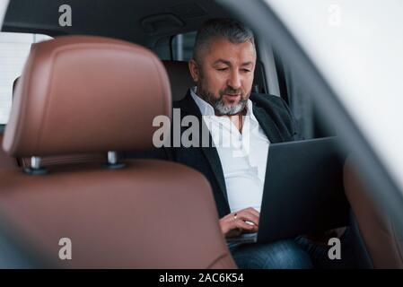 Bei Auftrag konzentriert. Arbeiten an einem Auto mit silbernen Laptop. Senior Geschäftsmann Stockfoto