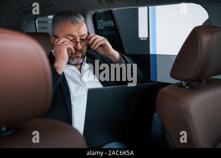 Das Setzen auf eine Brille. Arbeiten an einem Auto mit silbernen Laptop. Senior Geschäftsmann Stockfoto