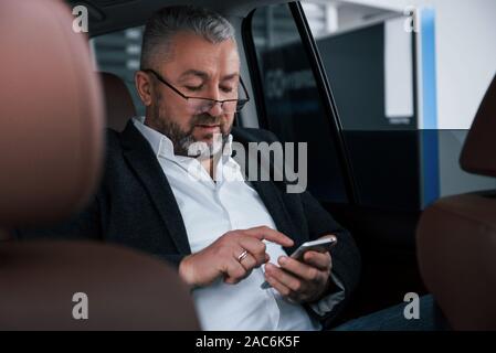 Neue Wege der Kommunikation ist die Arbeit leichter und schneller. Mit Handy. Unternehmer in der Brille sitzt auf der Rückseite des modernen Auto und Stockfoto
