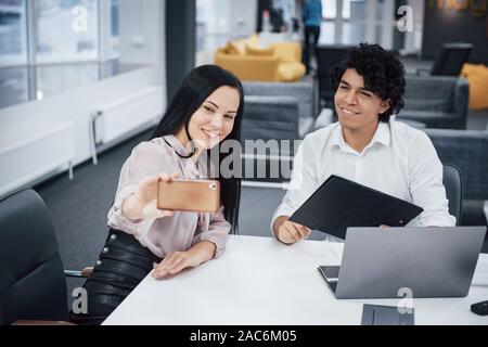 Selfie von zwei lächelnde Büroangestellte in offizielle Kleidung, die sitzt in der Nähe von Silver Laptop am Tisch Stockfoto