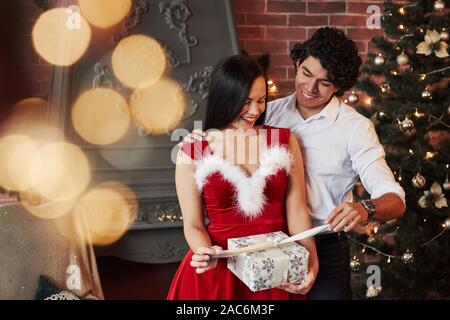 Eröffnung der Gegenwart. Schönes Paar feiern das neue Jahr in der dekorierten Zimmer mit Weihnachtsbaum und Kamin hinter Stockfoto