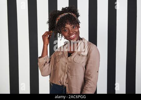 Lächelte afro-amerikanische Mädchen steht und berührt ihr Haar im Studio mit vertikalen weißen und schwarzen Linien im Hintergrund Stockfoto