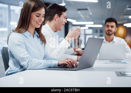 Frau geben Sie einen Text während der Kerl hinter Getränk aus Silber Glas haben. Seitliche Sicht auf Mädchen arbeitet auf der silbernen Laptop im Büro und lächelnd Stockfoto