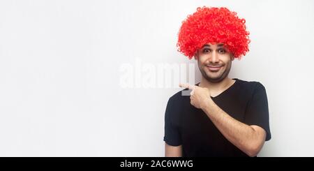 Schauen Sie hier! Portrait von stilvollen Lustig glücklicher Mann mit roten Perücke lächelnd und wies auf die Seite und zeigt leere Raum für Werbung, Freespace. Ich Stockfoto