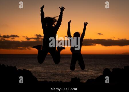 Silhouette von zwei junge Mädchen in der Nähe des Meeres im Sunset jugendlich Springen mit Ozean und orange Sonnenlicht im Hintergrund springen. Schwestern im Freien genießen. Stockfoto