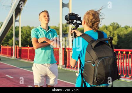 Junge teenager Modell und männlichen Fotografen Fotos und Videos mit der Kamera und Stabilisator, Dreharbeiten backstage, Hintergrund Fuß-Brücke über den Fluss auf Stockfoto