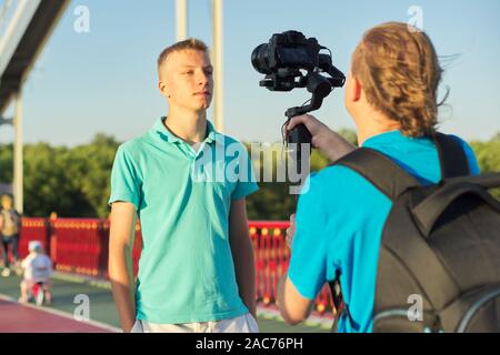 Junger Mann Modell und männliche Fahrer, Fotos Videos mit der Kamera und Stabilisator, Dreharbeiten backstage, Hintergrund Fuß-Brücke über den Fluss an sonnigen s Stockfoto