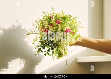 Schönen frischen Blumenstrauß von Maiglöckchen Blumen, Rosa, grüne Zweige in der Frau die Hand, Hintergrund Licht sonnige Wand. Floristik, Arbeit von floris Stockfoto