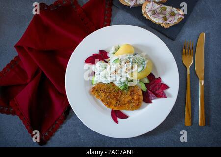 Essen Fotografie eines vegane Schnitzel, gekochte Kartoffeln und eine Dill und Joghurt Sauce auf einem schwarzen Schiefer Hintergrund und ein rotes Tuch Stockfoto