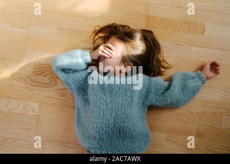 Kleines Mädchen mit einem blauen Pullover auf dem Boden liegend, die Augen vor Sonnenlicht Stockfoto