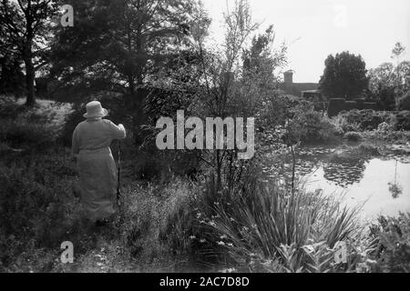 Alte Dame in einem feinen Frock und Hut am Horse Pond, mit Christopher Lloyd's Garten in Great Dixter, East Sussex. Altes Schwarzweiß-Filmfoto, ca. 1980 Stockfoto