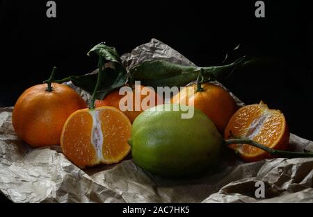 Tangerine ganze und Open-face mit Blatt und eine grüne Zitrone auf Küchenpapier auf schwarzem Hintergrund Stockfoto