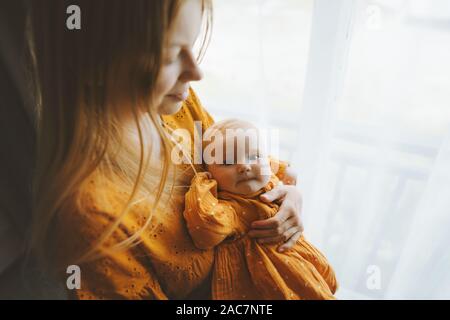 Mutter Holding baby Tochter Familie lifestyle Mutter und Kind zusammen zu Hause Mutterschaft Konzept Muttertag urlaub Senf Farbe Kleid Stockfoto