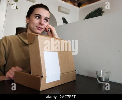 Ein junges Mädchen sitzt an einem Tisch in einem Cafe und schaut weg mit einem verträumten Blick. Vor der Mädchen ist ein offenes Feld mit einem Geschenk. Für Text platzieren. Stockfoto