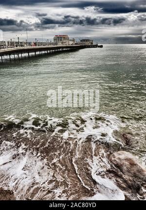 Portraitfotos der Wellen gegen die Küste mit viktorianischen Worthing's Pier im Vordergrund und Rapunzeln Offshore Windpark am Horizont Stockfoto