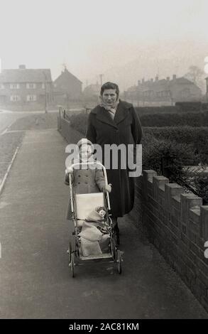 1950, historische, eine Mutter in Mantel und Schal, verpackt in einer Wohnsiedlung mit ihrer jungen Tochter, drückt eine kleine Kinderwagen oder Buggy der Ära mit ihren kleinen Puppe, die mit einer Decke, England, UK gewickelt ist. Stockfoto