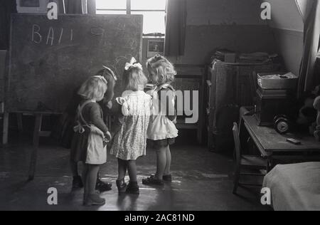 1950, historische, Nachkriegszeit, Großbritannien und in einem Zimmer in einem Kindergarten, kleine Mädchen in Schürzen zusammen an einer Tafel stehen. Eine große Anstrengung, war nach dem Krieg Vorschule lernen Raum für die frühkindliche Bildung zu bieten, bevor sie der Schulpflicht in der Grundschule beginnen. Stockfoto