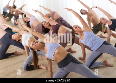 Gruppe von jungen sportlich attraktive Leute im Yoga Studio, Yoga Unterricht mit Kursleiter, Stretching auf Fußboden im Trikonosana, Dreieck yoga Pose Stockfoto
